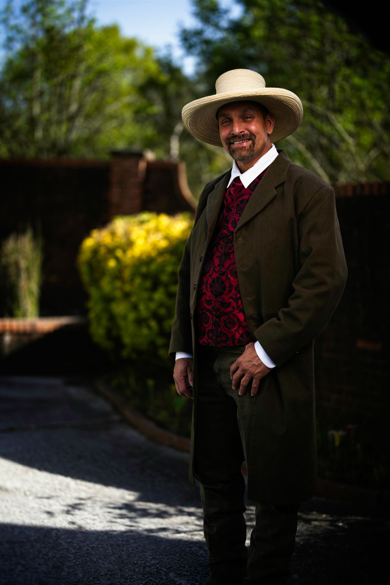 A man in a suit and hat is standing in front of a bush.