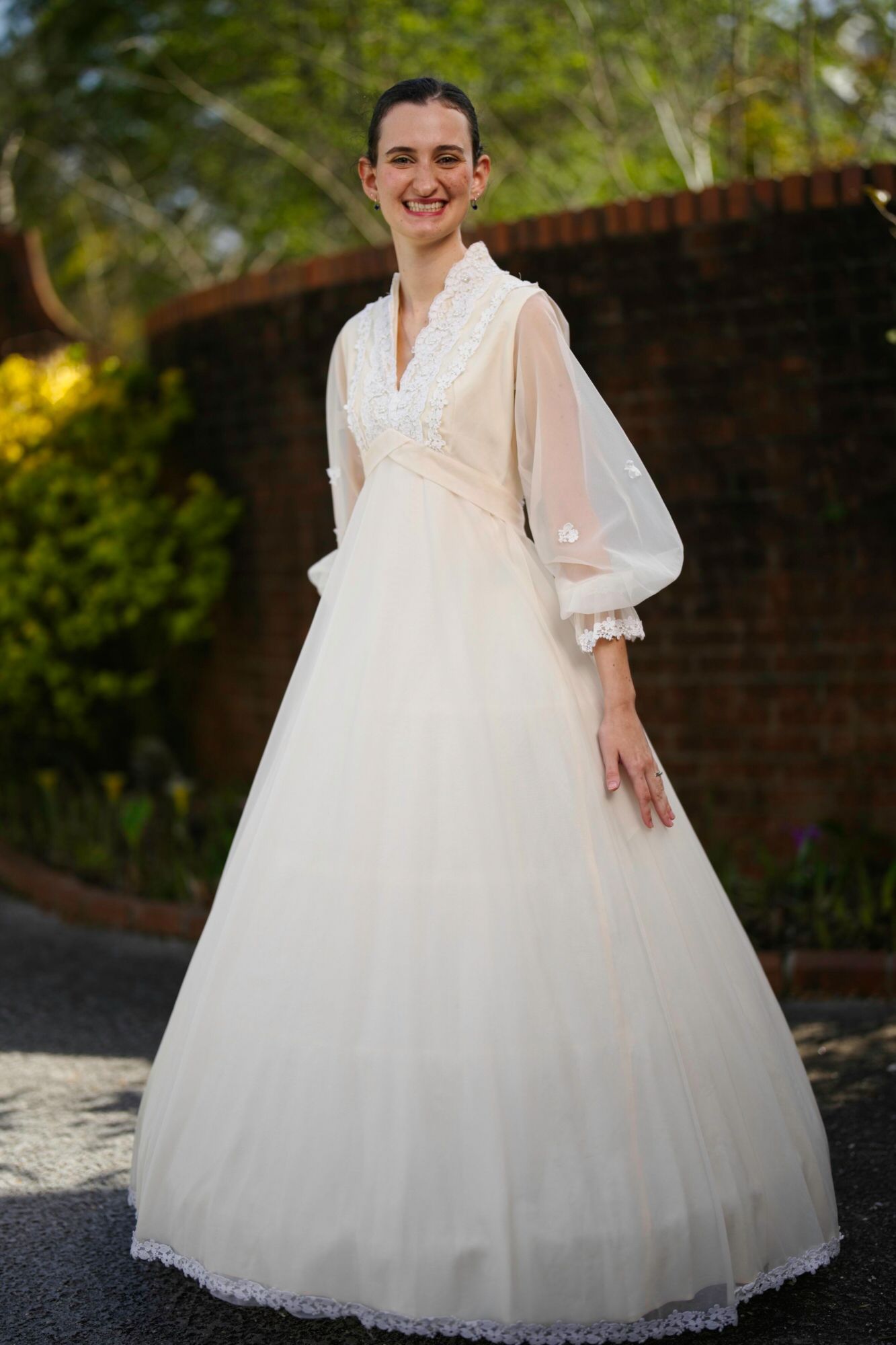 A woman in a white dress is standing in front of a brick wall.