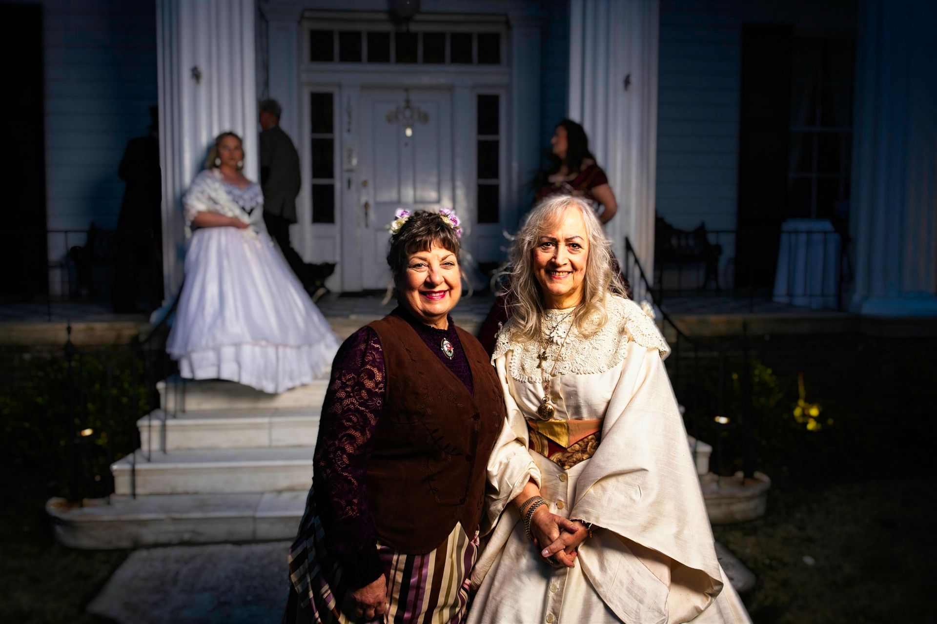 Two women in historical costumes are posing for a picture in front of a house.