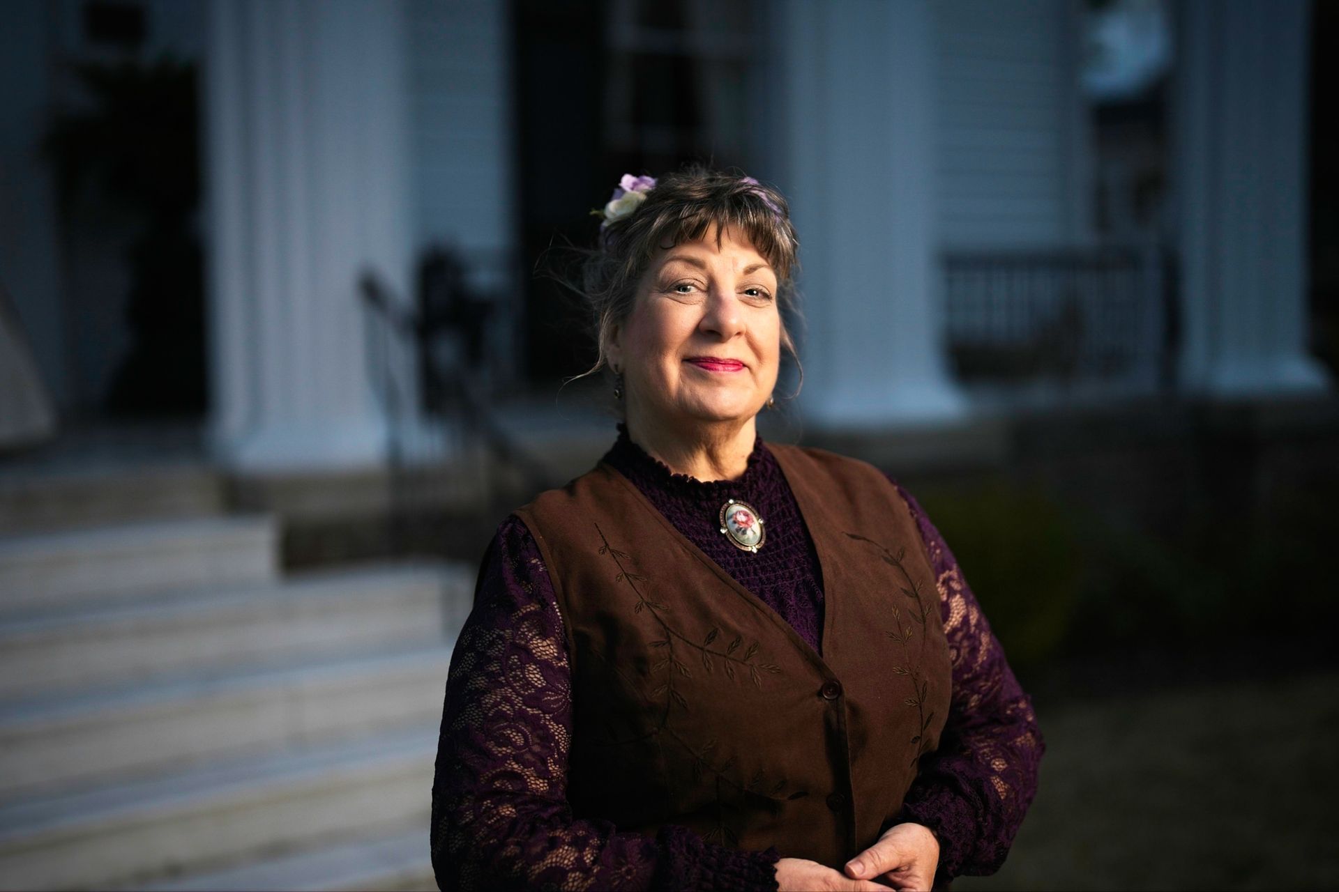 A woman in a brown vest is standing in front of a house.