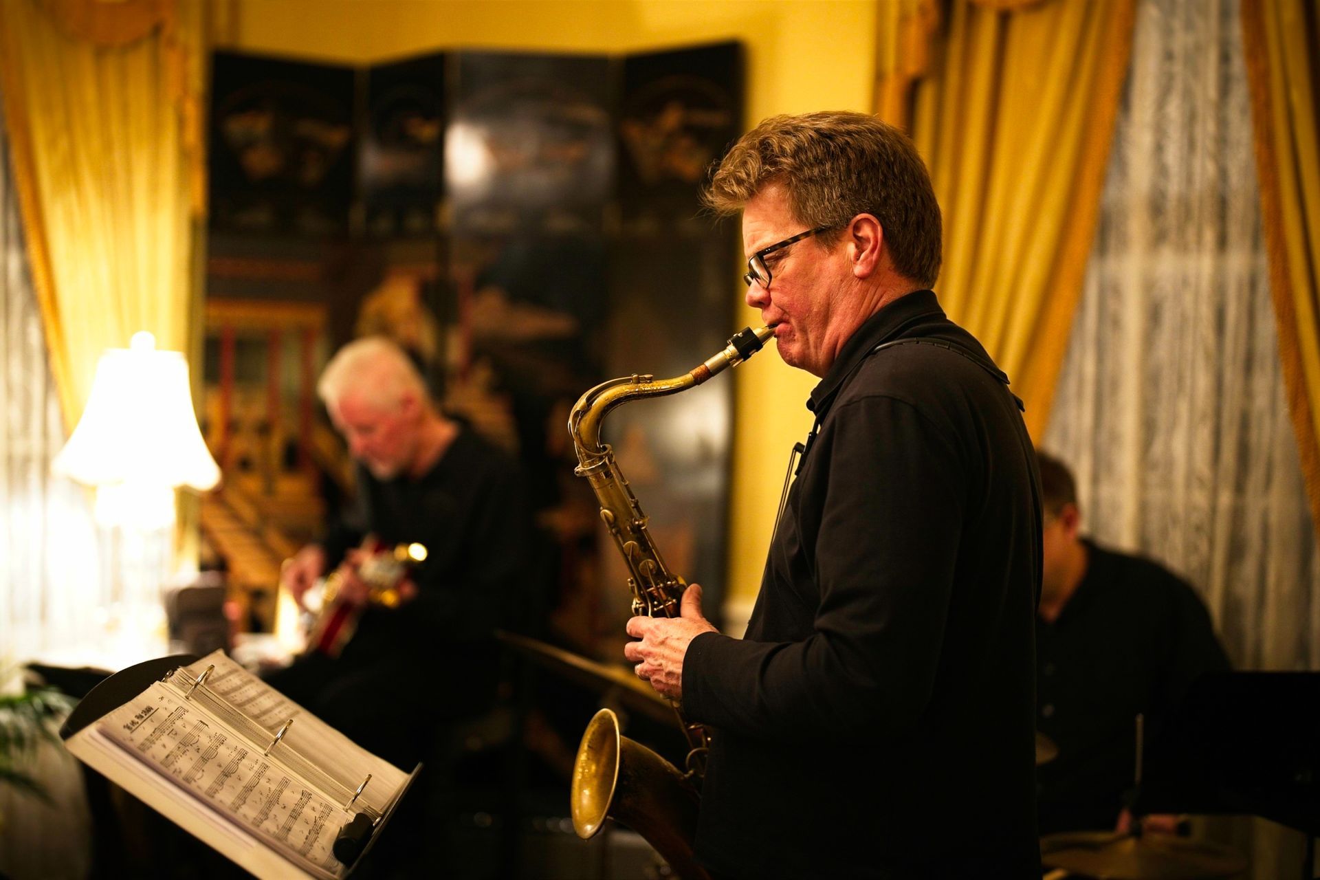A man is playing a saxophone in a room while another man plays a guitar.