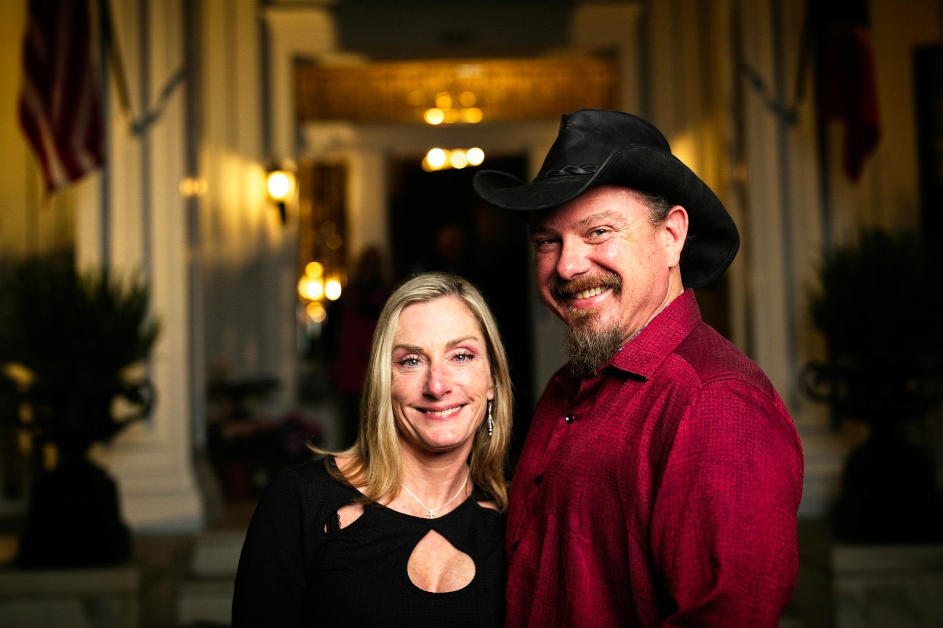 A man and a woman are posing for a picture in front of a building . the man is wearing a cowboy hat.
