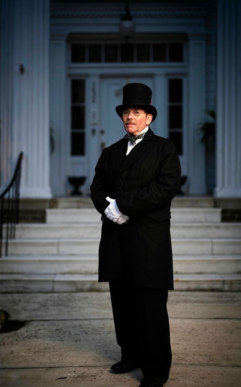 A man in a top hat and coat is standing in front of a building.