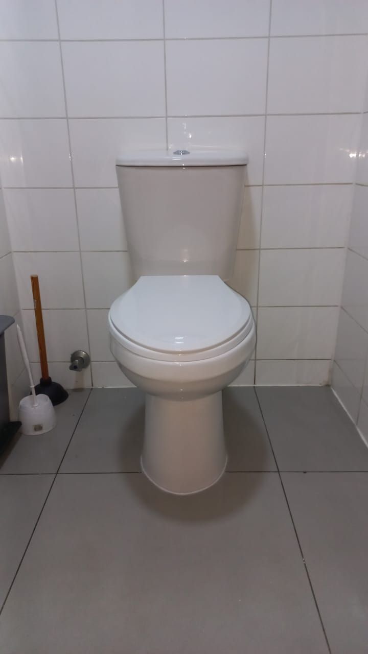 Inodoro blanco en un baño con suelo gris y paredes de azulejos blancos. Hay una escobilla de baño cerca.