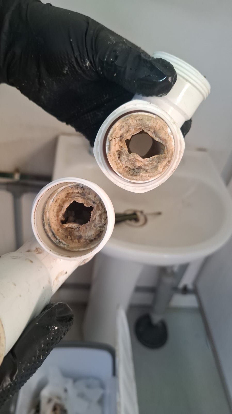 Dos tubos de plomería de plástico blanco, muy calcificados, sostenidos por manos enguantadas en negro cerca de un fregadero.