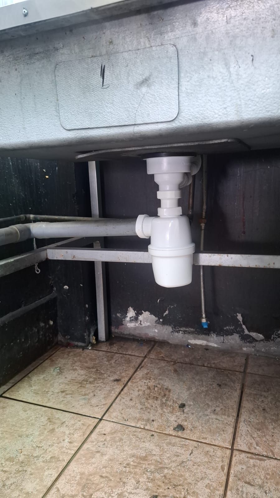 Parte inferior de un fregadero de cocina: sifón de plástico blanco y tuberías sobre un fondo oscuro y un suelo de baldosas.