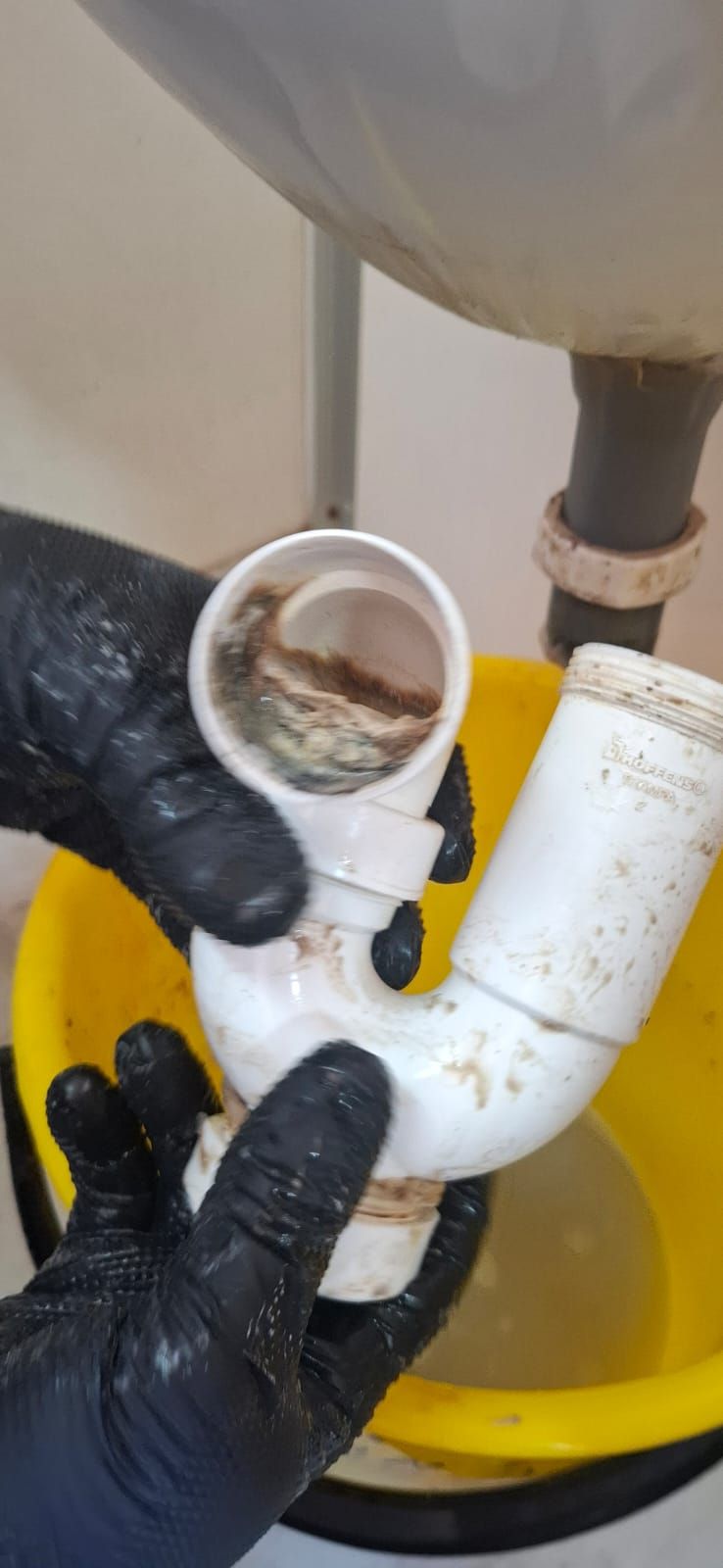 Manos con guantes negros que sostienen un sifón de plomería blanco desmontado y cubierto de suciedad y escombros.