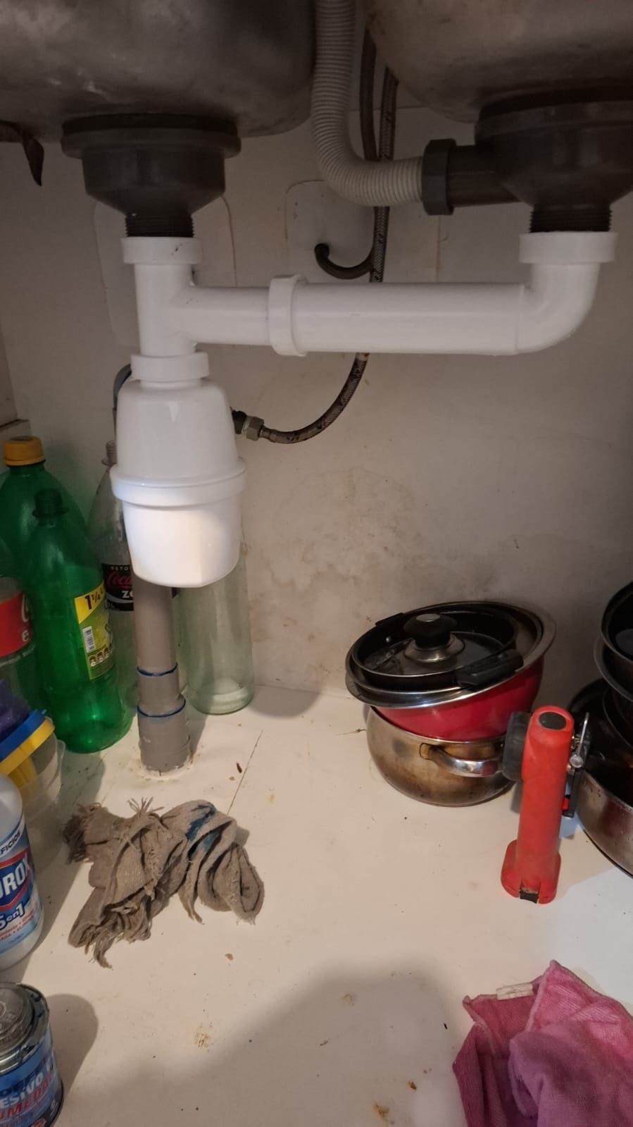 Tuberías de desagüe de fregadero de cocina blancas debajo de un fregadero, con desorden en el estante de abajo.