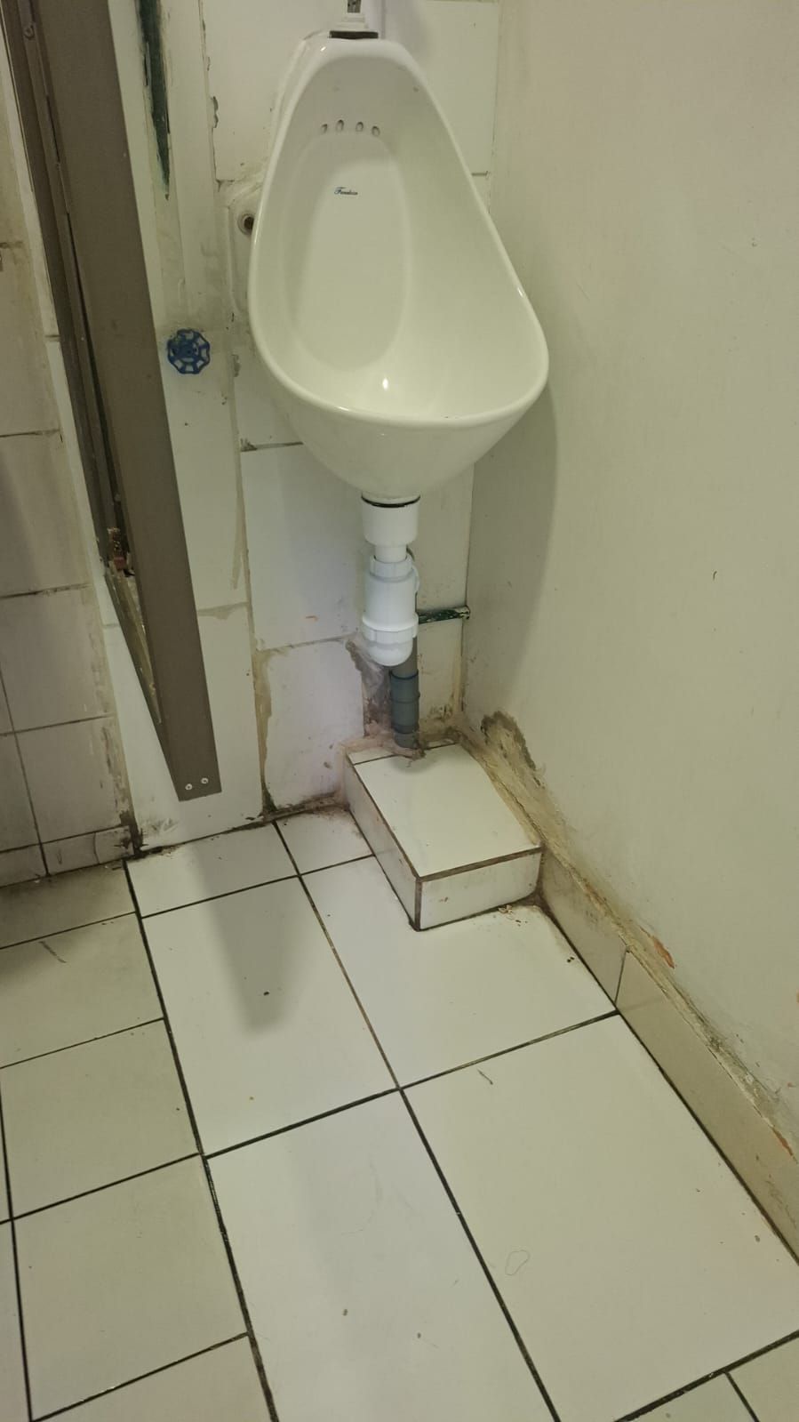 Urinario en una esquina, sobre un pequeño escalón. Cerámica blanca, tubería gris y baldosas blancas y sucias.