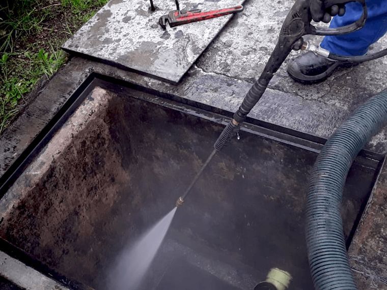 Una persona usa una hidrolavadora para limpiar una abertura cuadrada oscura. Sale agua a chorro. Hay una llave inglesa y una manguera cerca.