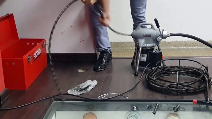Persona que utiliza una máquina de limpieza de desagües en el suelo; caja de herramientas y cable de limpieza de desagües visibles.