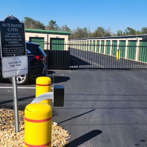 An automatic gate at a storage facility with a black car parked near the entrance. Green storage units are visible in the background.