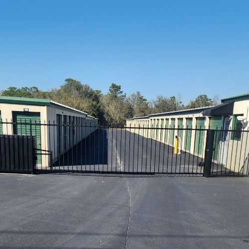 Storage units behind a black gate on an asphalt lot, under a clear blue sky.