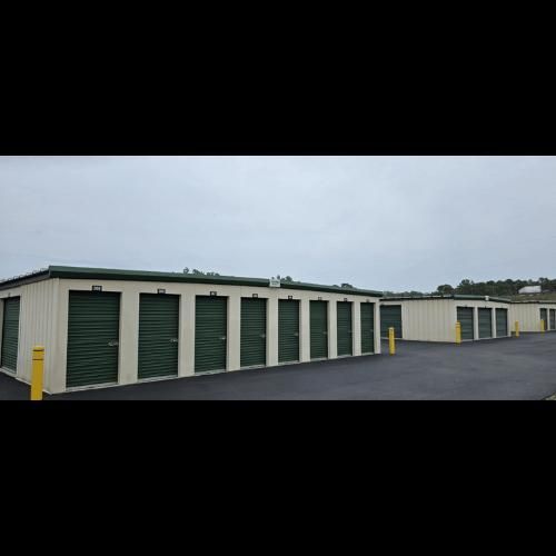 Storage units with green doors in a row, on a paved area, under an overcast sky.