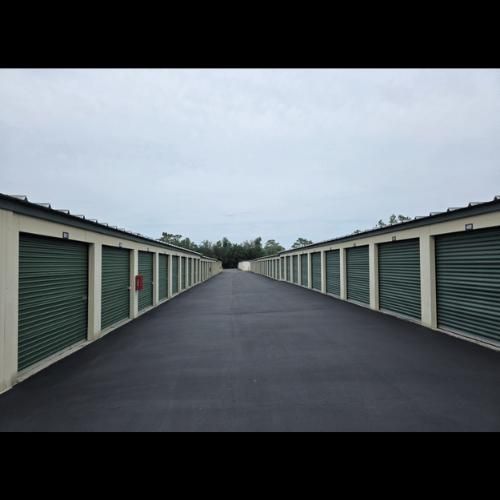 Rows of green storage unit doors line a long asphalt drive under a cloudy sky.