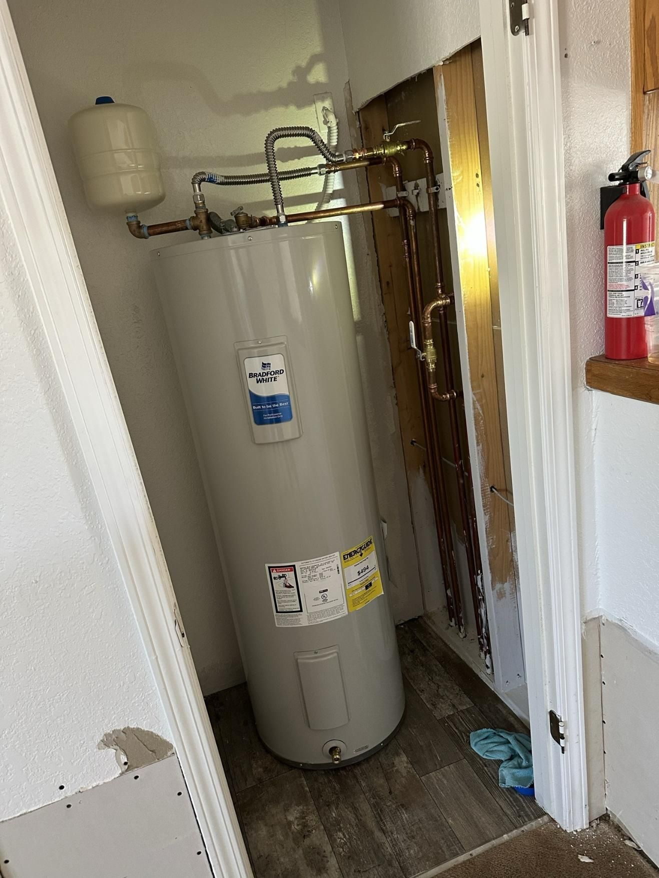 Water heater in a closet with a white expansion tank. Brown wood paneling. A fire extinguisher hangs on the wall.