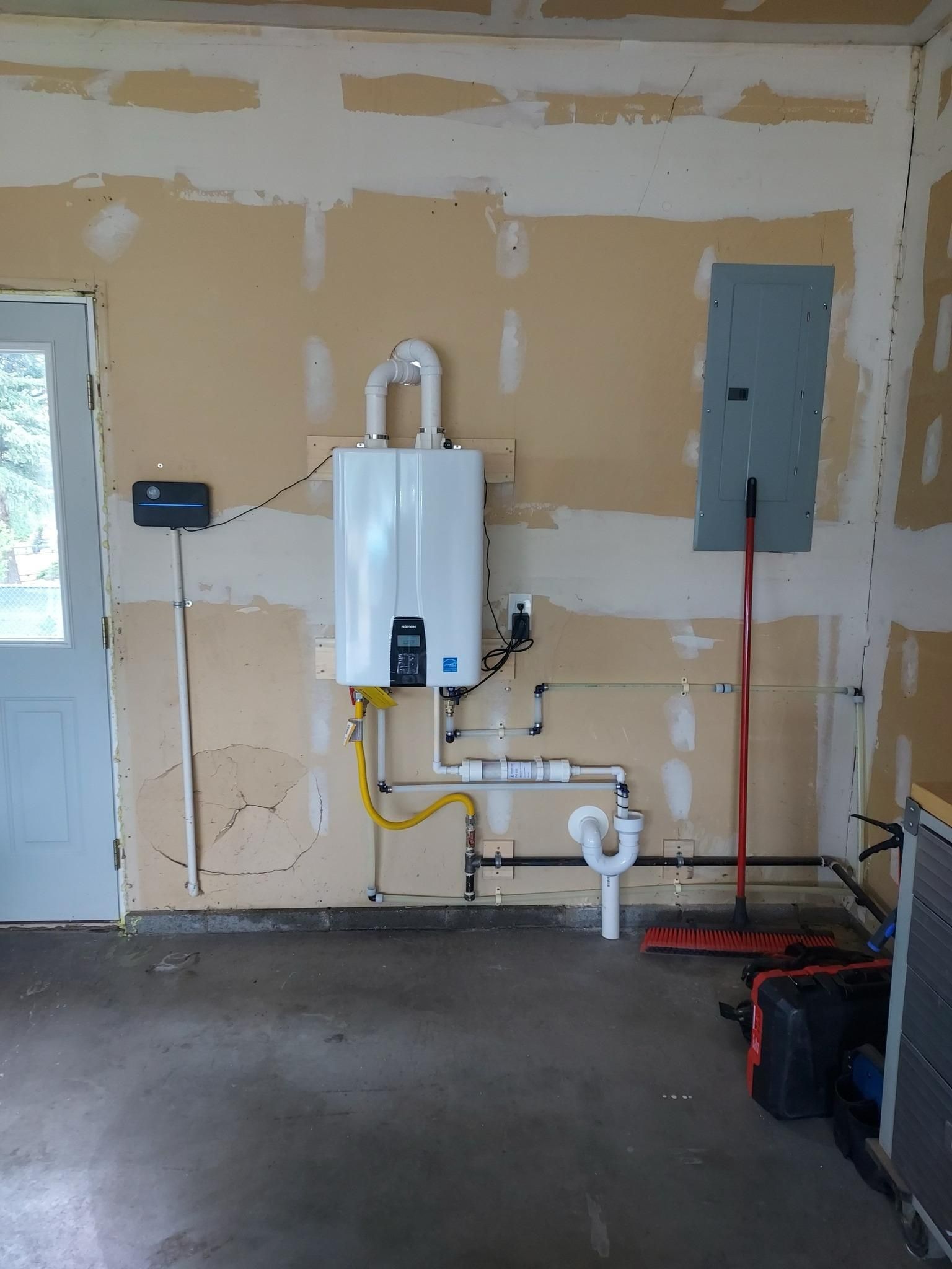 White tankless water heater installed in a garage, next to a breaker box and plumbing.