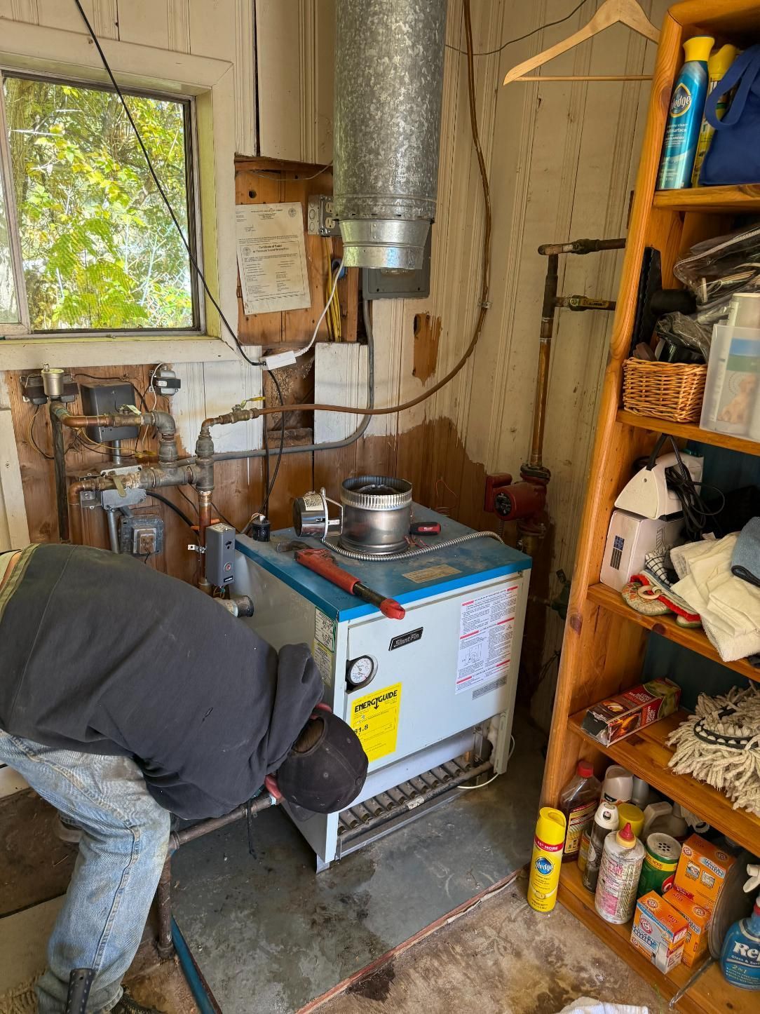 A person repairs a blue and white water heater in a cluttered utility room.
