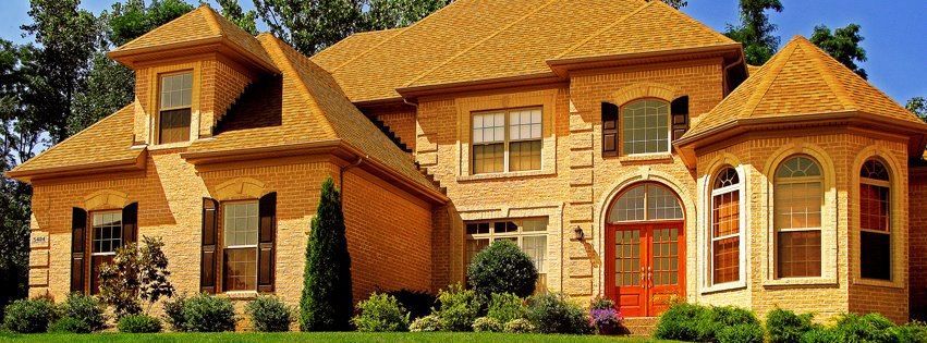 Large, brick house with a brown roof and shutters, a red front door, and manicured landscaping.