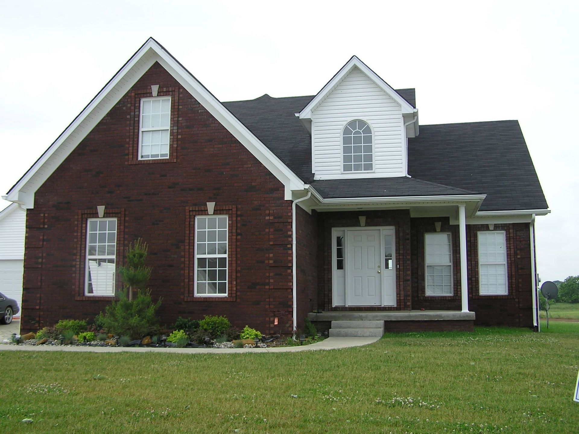 Red brick house with white trim, black roof, and front porch on a green lawn.