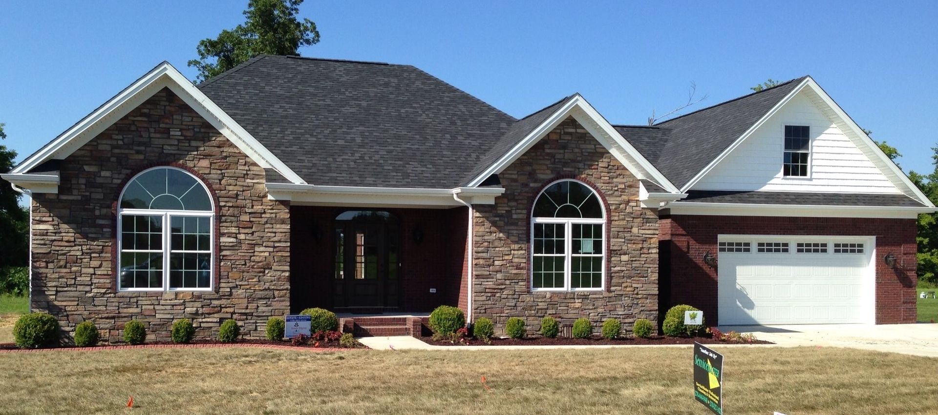 House with stone and brick exterior, dark roof, arched windows, and white garage door under a blue sky.