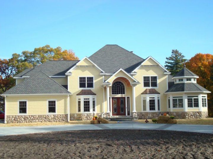 Yellow two-story house with stone base, gray roof, arched front door, and circular side room.