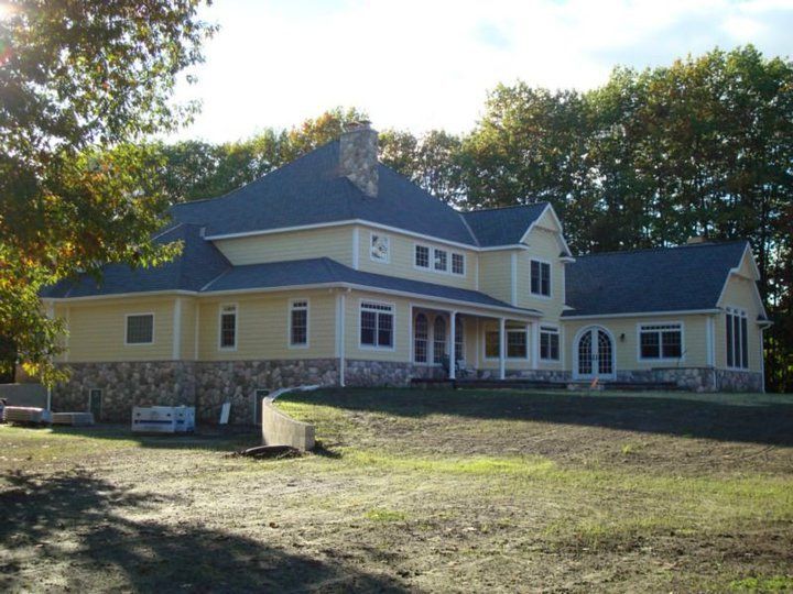 Yellow house with dark roof and stone base, set on grassy hill, trees in the background.