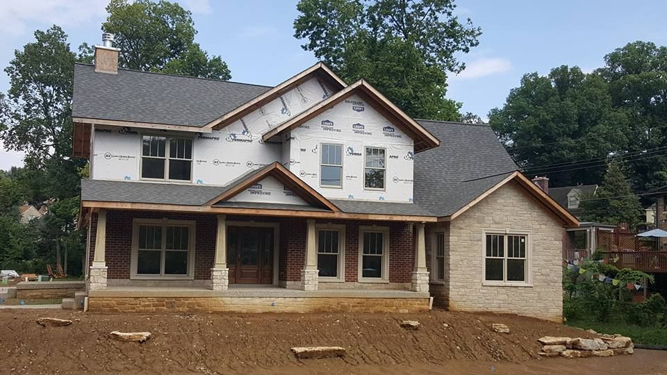 A two-story house under construction with stone and brick exterior features, surrounded by dirt and trees.