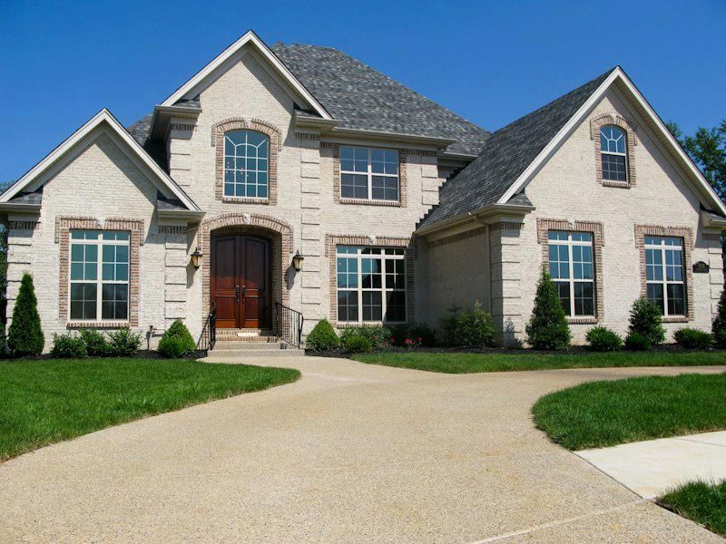 Two-story beige brick house with gray roof, arched front door, and a circular driveway.