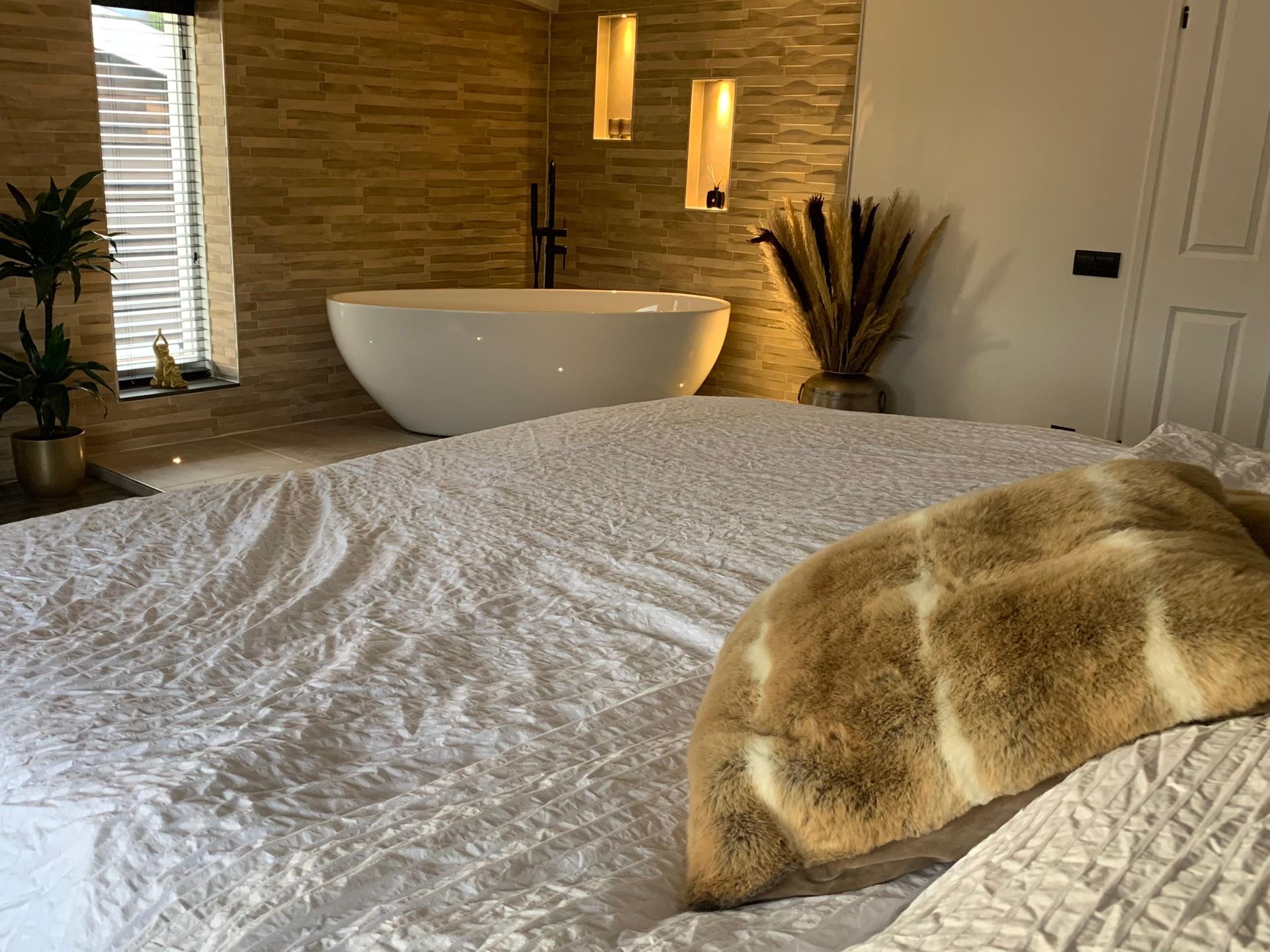 Een wit ovaal bad in een slaapkamer met muren van natuursteen, warme verlichting en een beige sierkussen met textuur op het bed.