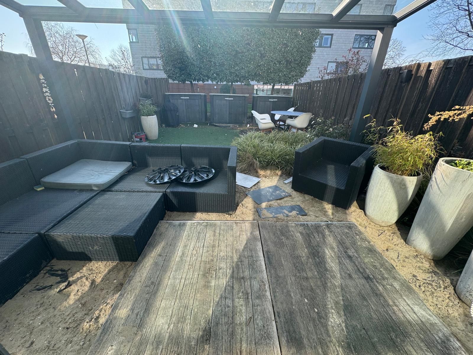 Buitenpatio met een donkere rieten bank, een bijpassende fauteuil, houten planken op zand en grote witte plantenbakken.