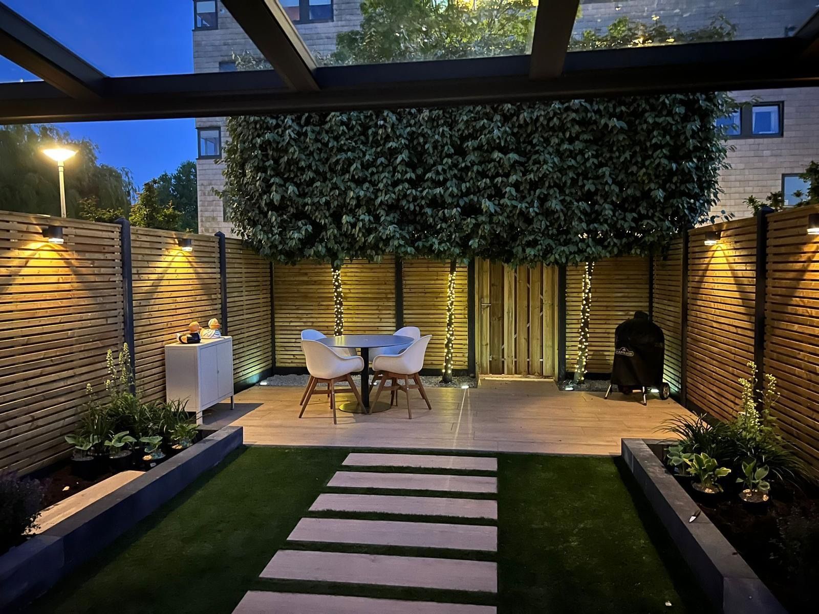 Gezellig terras met zitgelegenheid, groen en sfeerverlichting. Een pad leidt naar een eethoek onder een afdak.