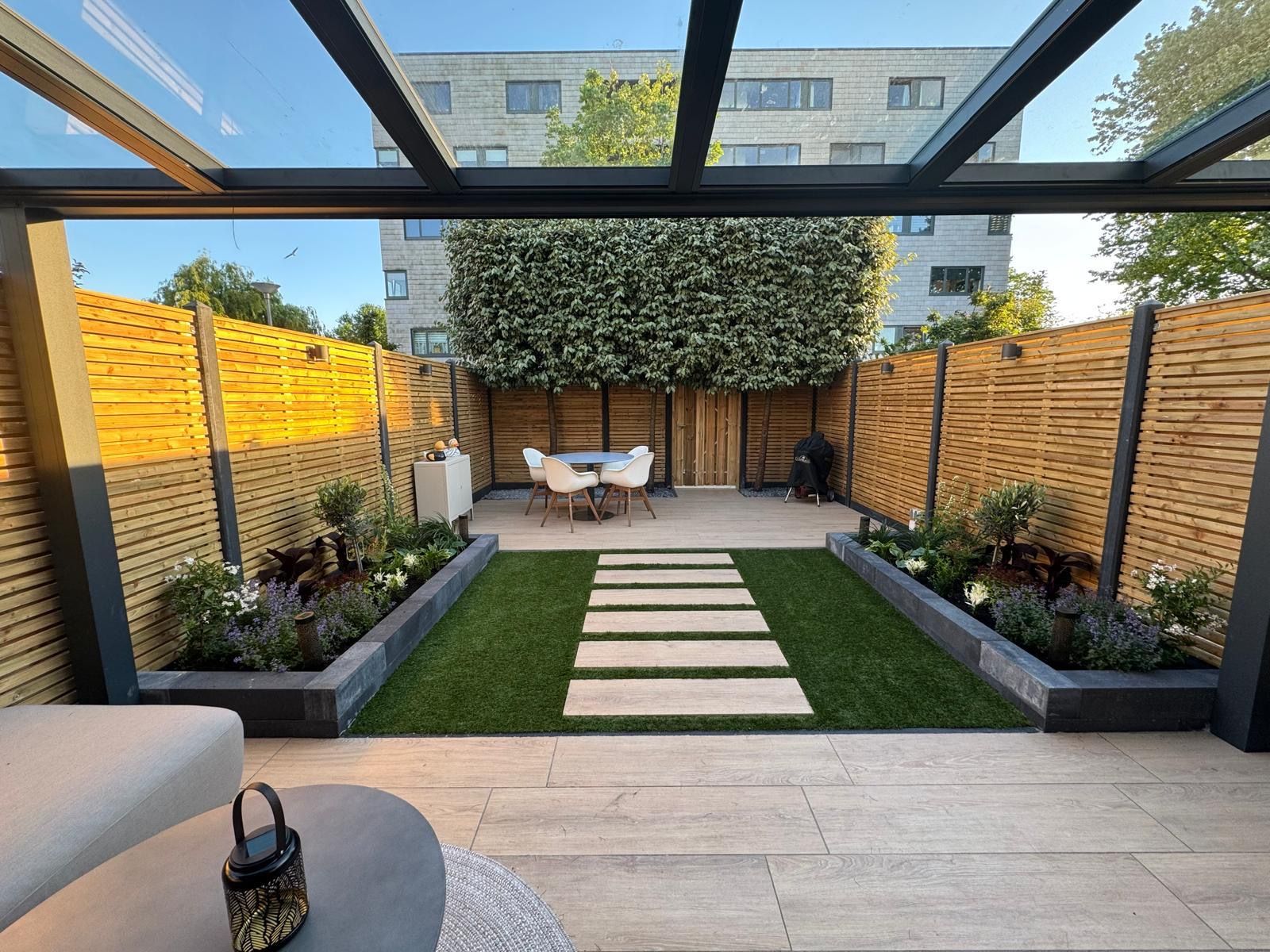 Een moderne achtertuin met een glazen dak, een centraal graspad met natuurstenen bestrating en een houten schutting eromheen.