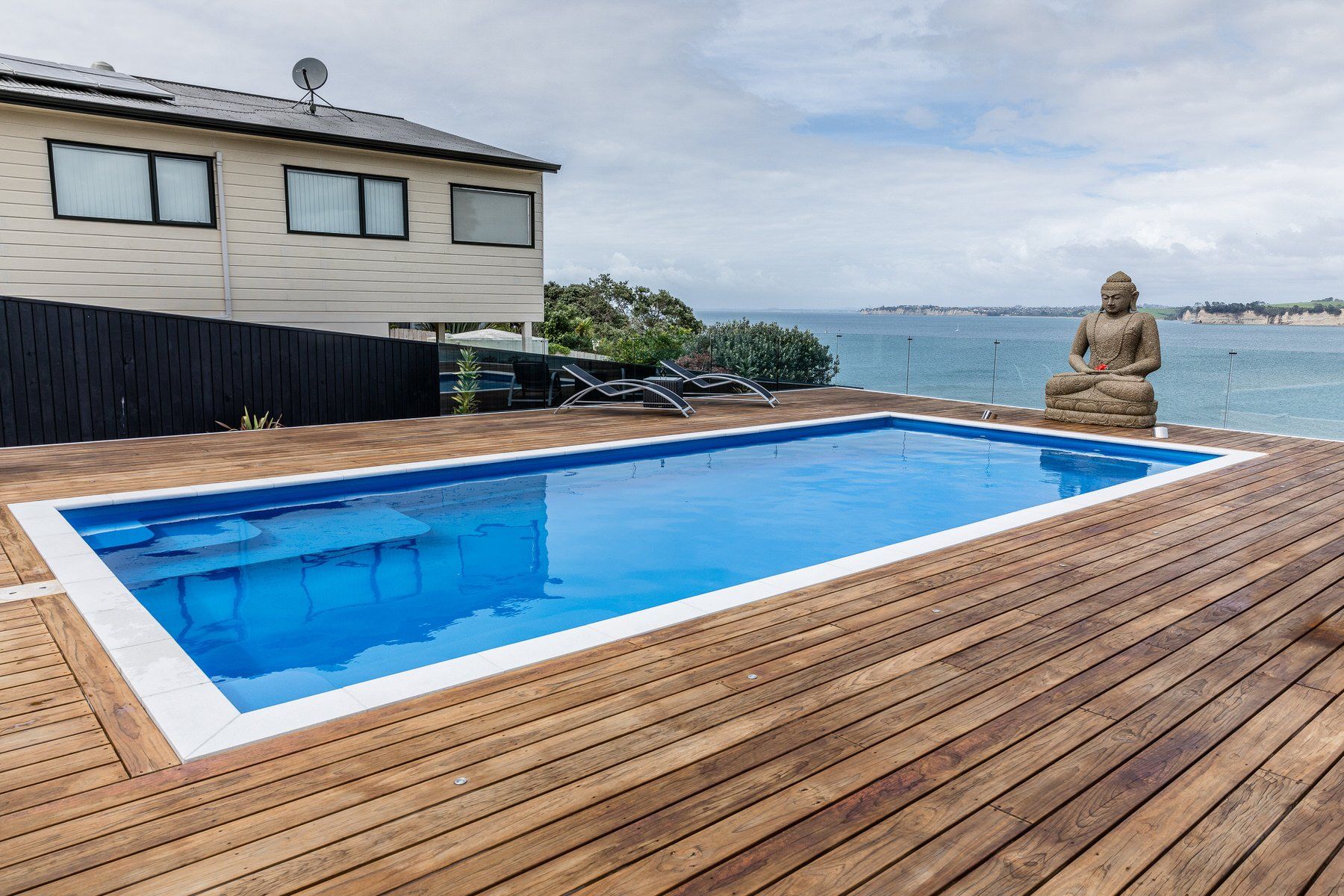 A large swimming pool is sitting on top of a wooden deck next to the ocean. Poolcorp Pools Auckalnd