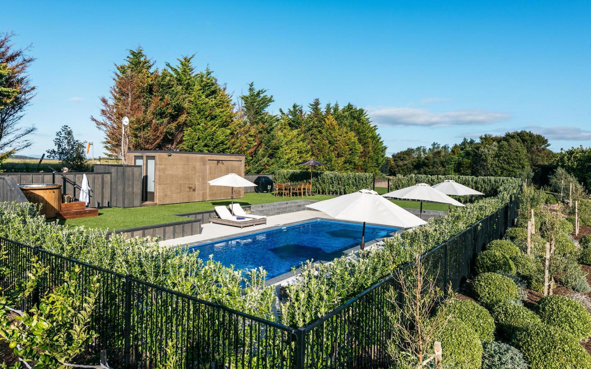 A large swimming pool surrounded by a fence with umbrellas and chairs. Poolcorp Pools Auckland