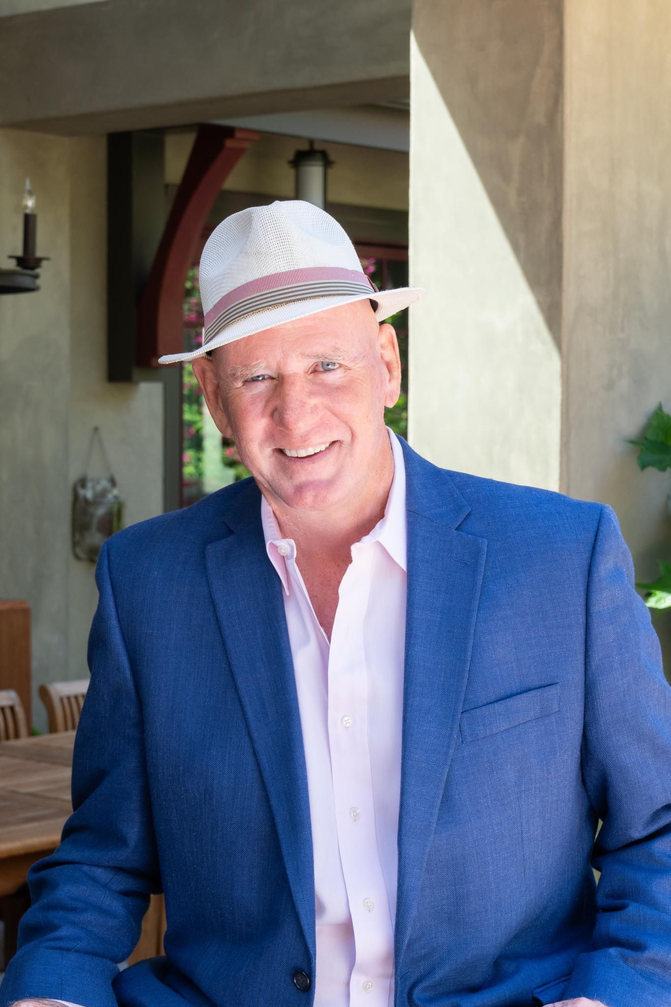 A man wearing a blue suit and a white hat is smiling.