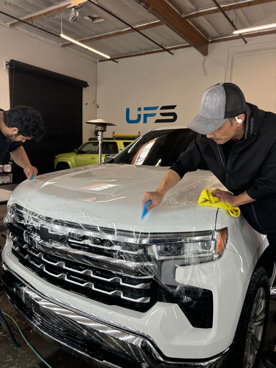 Two people detail a white SUV in a workshop, wiping the hood and front grille.