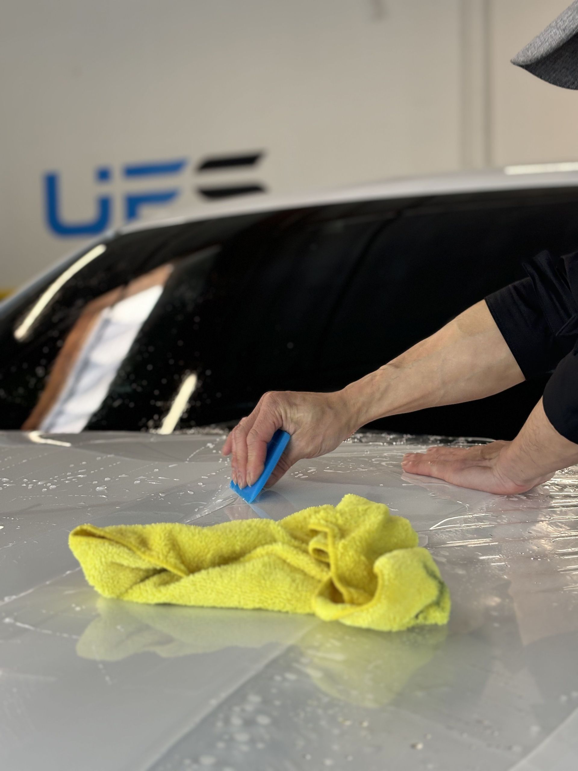 Hand polishing a car hood with a yellow cloth and blue applicator in a garage