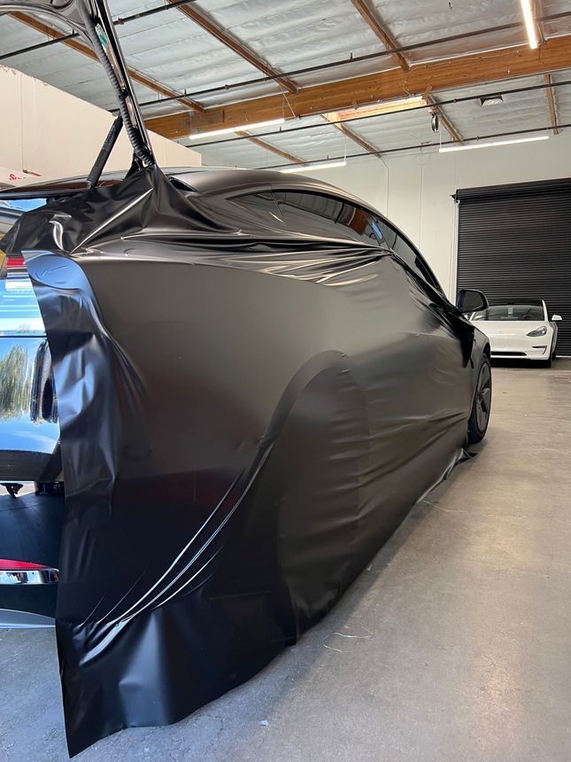Black car covered in a glossy protective wrap inside a garage with exposed ceiling beams