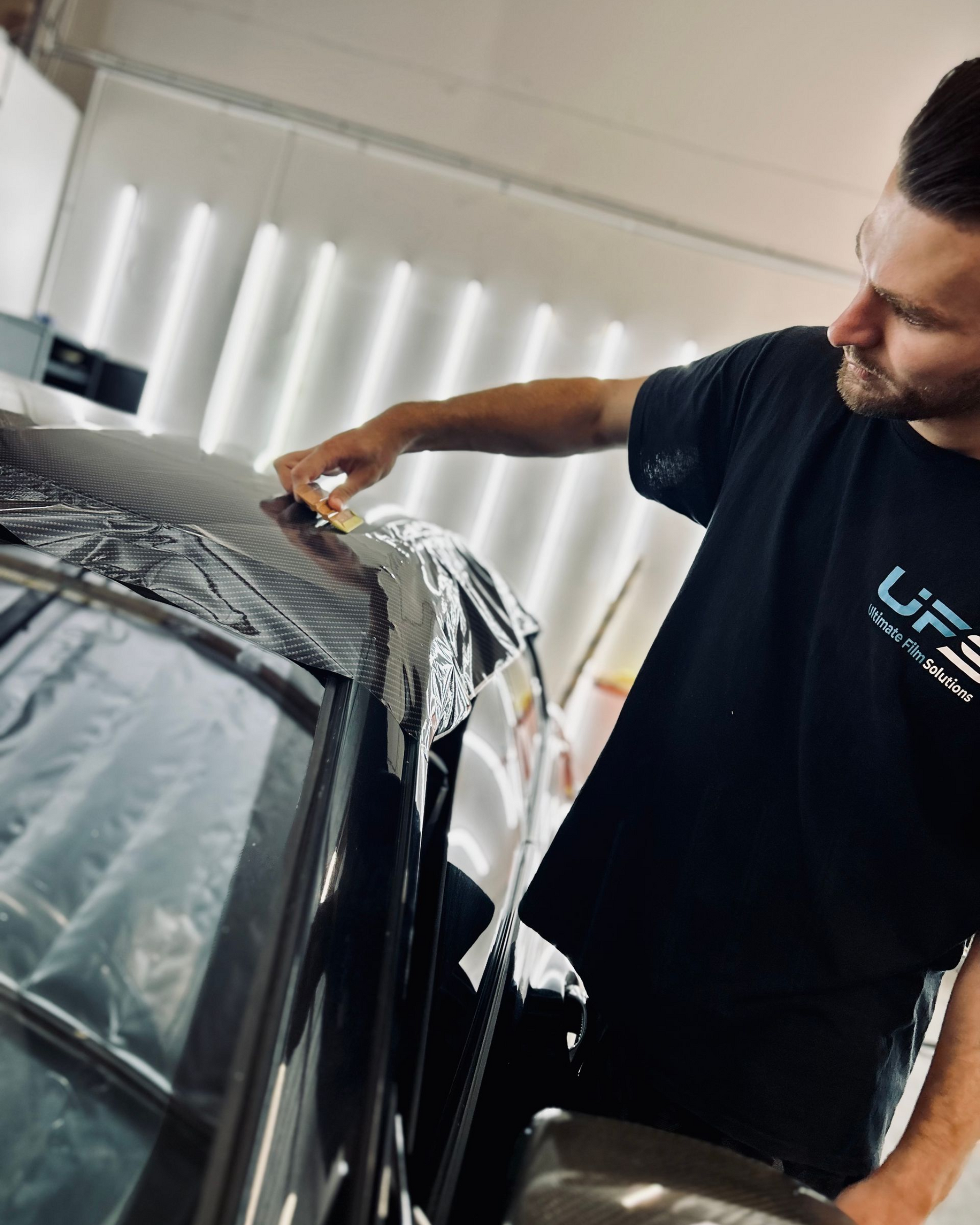 Man applying vinyl wrap to a black car indoors, leaning over the roof with tools nearby