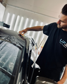 Man applying vinyl wrap to a black car indoors, leaning over the roof with tools nearby