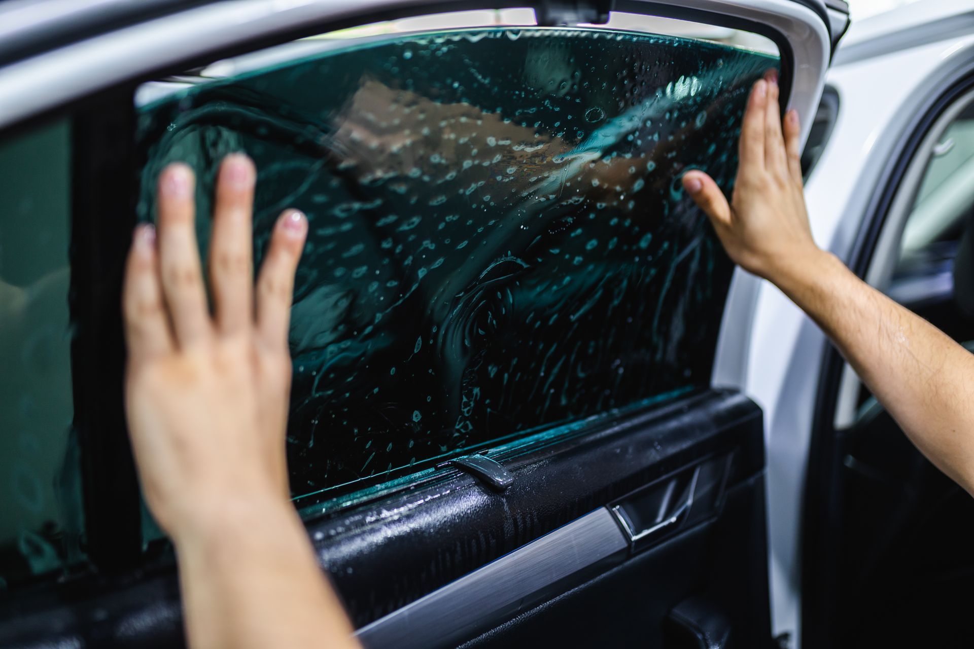Hands peeling tint from a car window, with water droplets on the glass