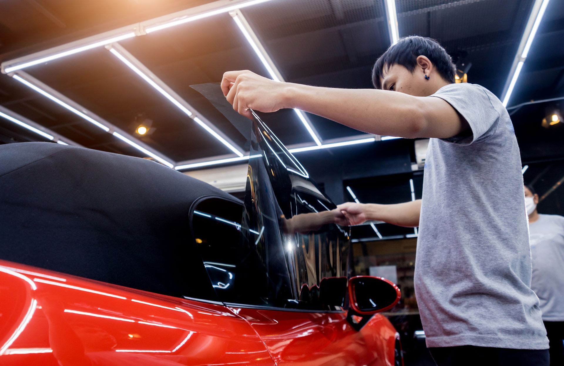 Person applying film to a red car window in a workshop with bright ceiling lights