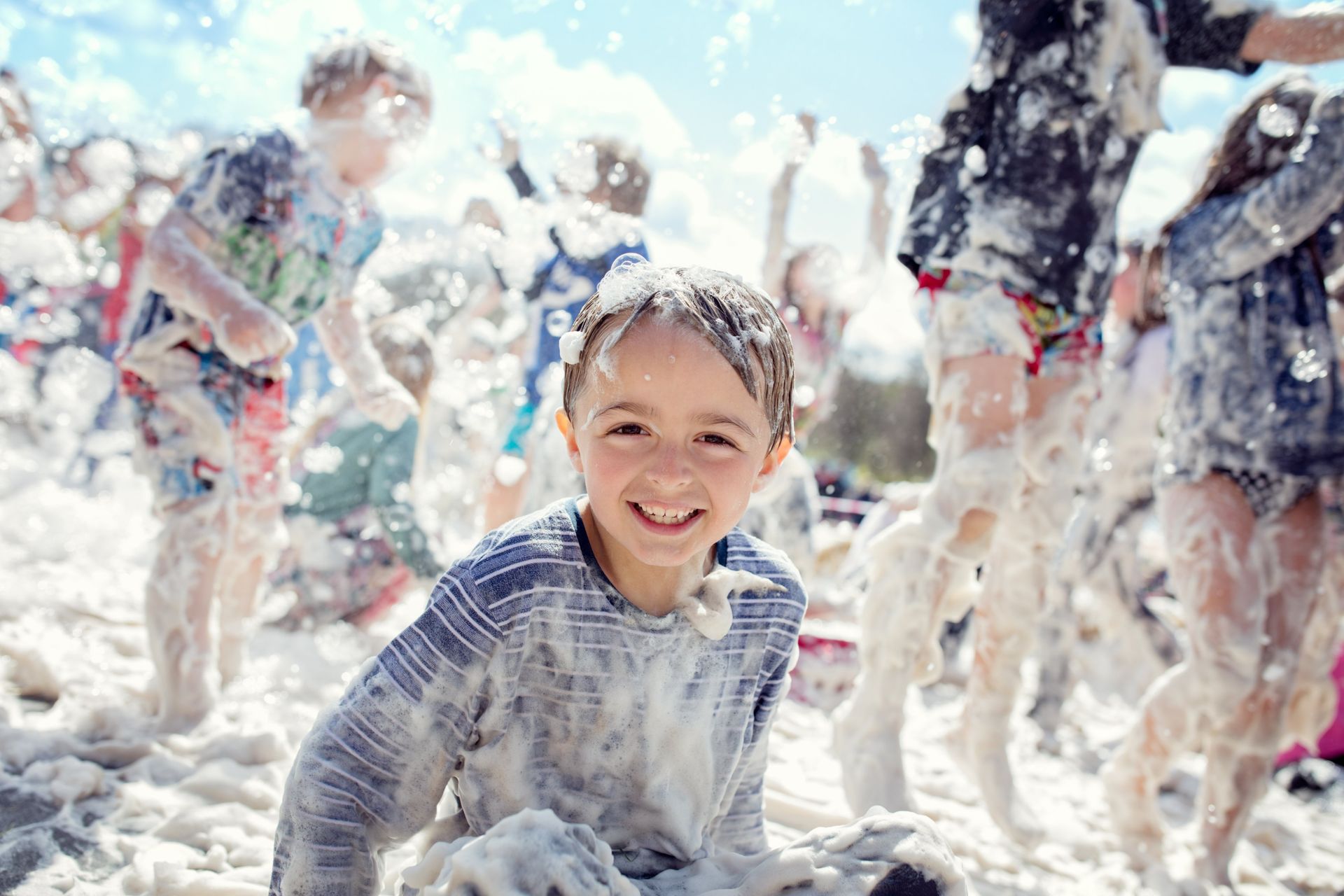 Boy smiles, covered in foam, amid other children in a foam party.