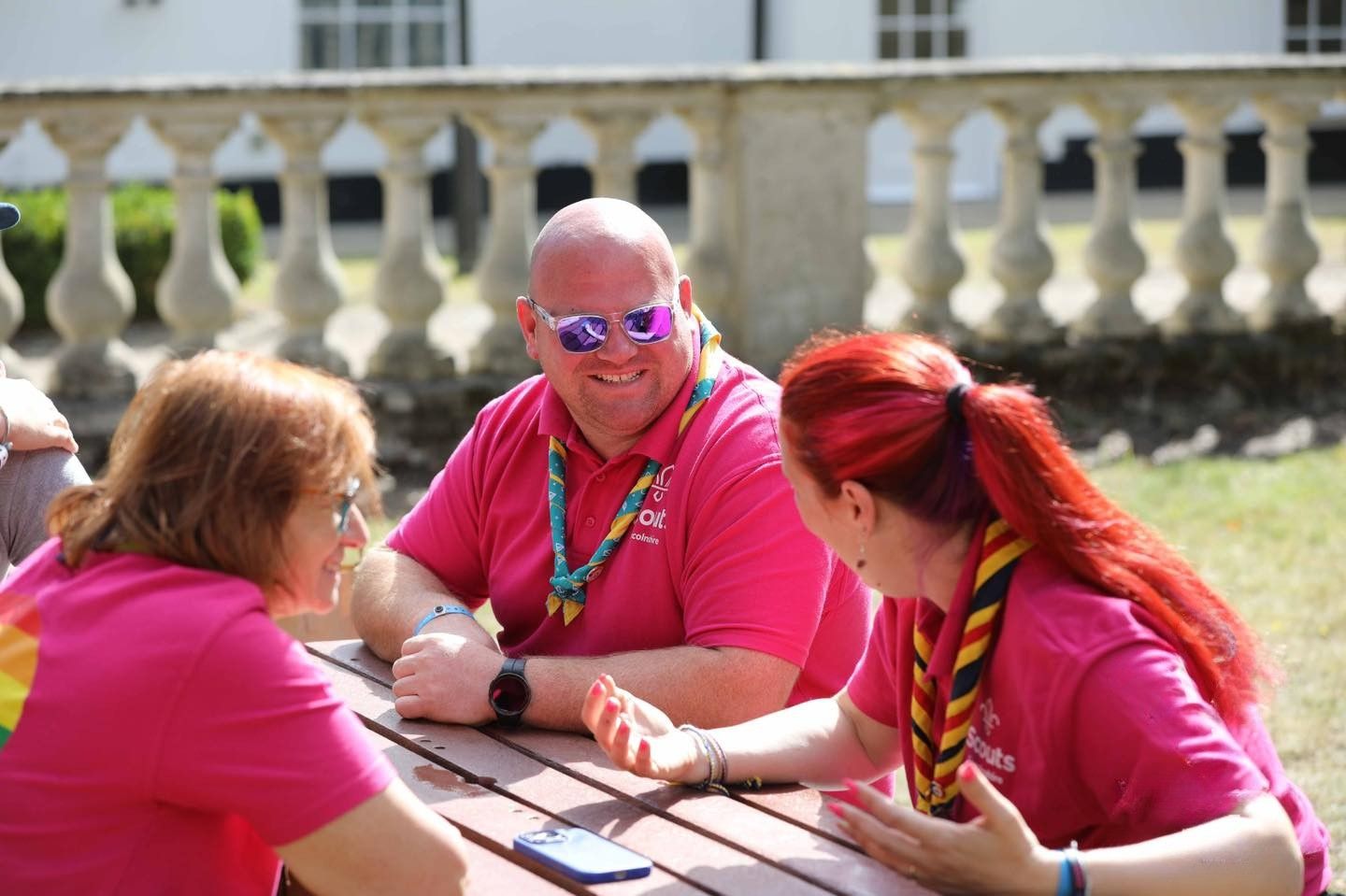 District Scout volunteers sitting around a picnic bench chatting and smiling, wearing bright pink Scouting Pride T-shirts, showing teamwork and inclusion in action.