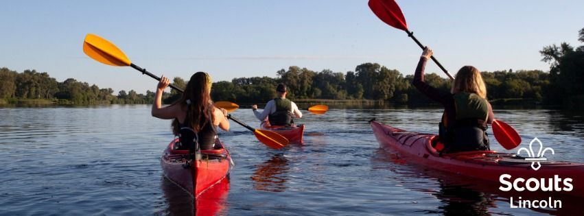 Three young people kayaking on a calm lake with paddles raised, part of Lincoln Scouts water activities.