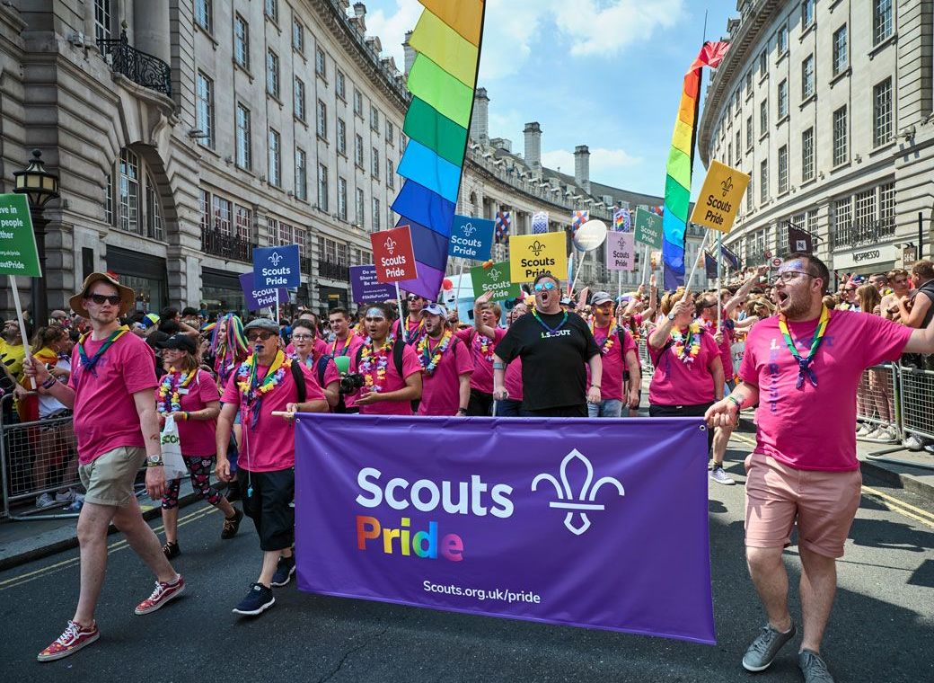 Scouts Pride group marching at London Pride, wearing pink shirts and rainbow accessories, carrying a large purple Scouts Pride banner and rainbow flags through a busy city street.