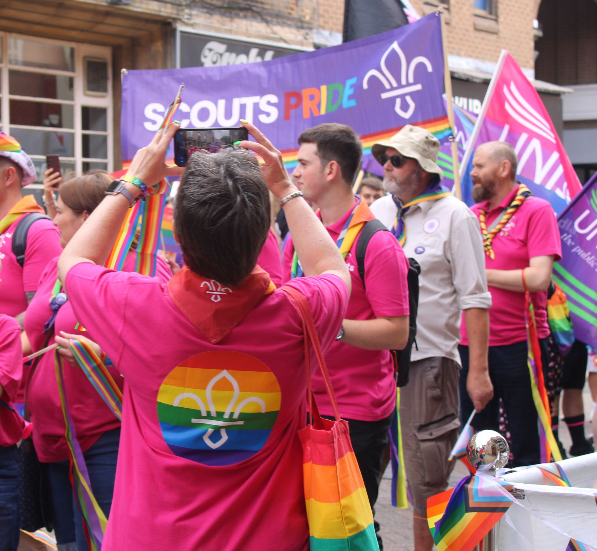 Lincoln Scouts members at Pride wearing pink shirts with rainbow Scout logos, standing in front of a Scouts Pride banner and rainbow flags.