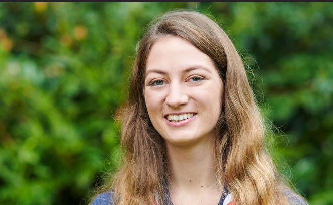 Smiling Scout volunteer with long hair standing outdoors in front of greenery.