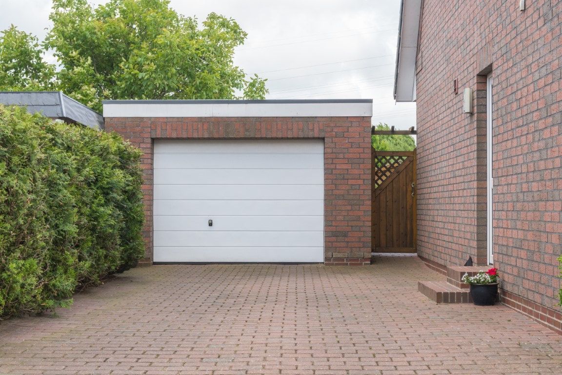 Brick garage with white door at the end of a paved driveway, bordered by hedges and a side wall.